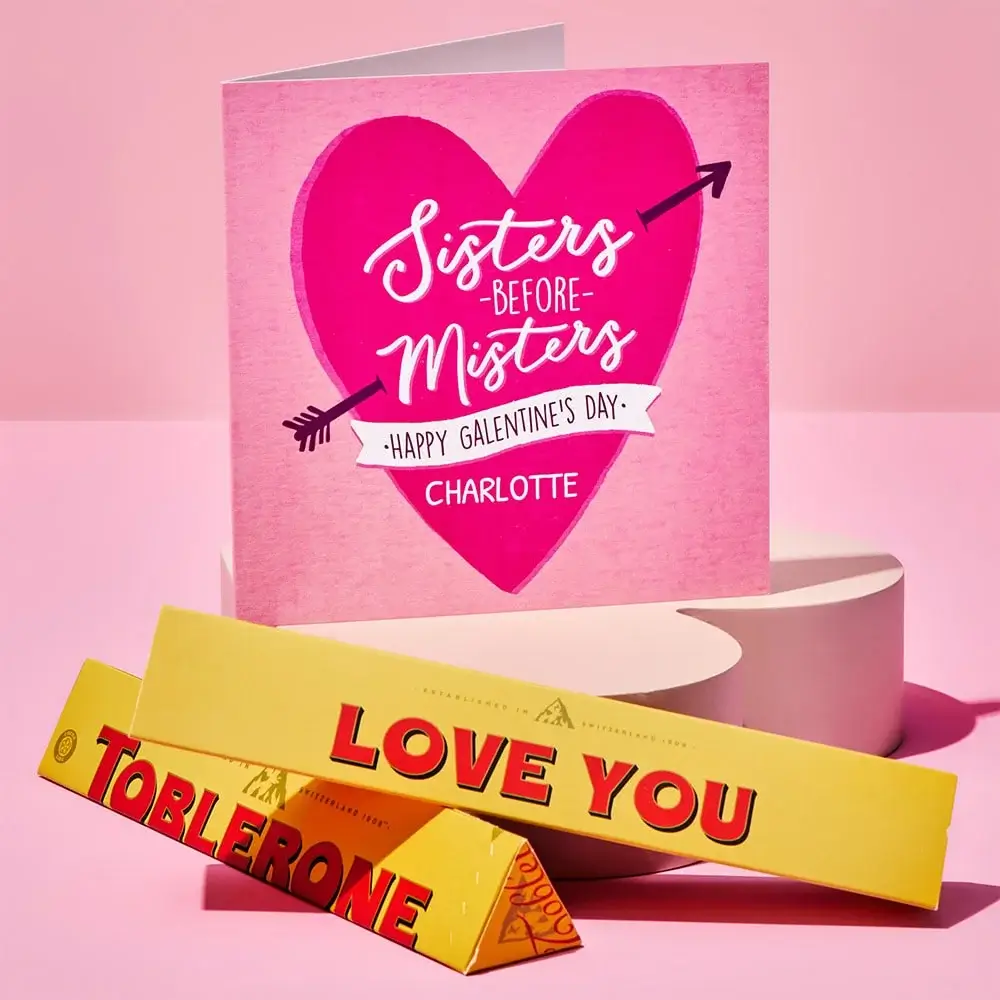 Pink Galentine’s Day card with a large heart and arrow design reading Sisters Before Misters, placed behind two yellow Toblerone chocolate bars with Love You text, set on a pink background.